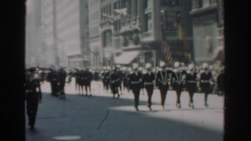 Uniformed marchers continue through a New York City street scene, with a crowd behind them and an American flag at the right edge.