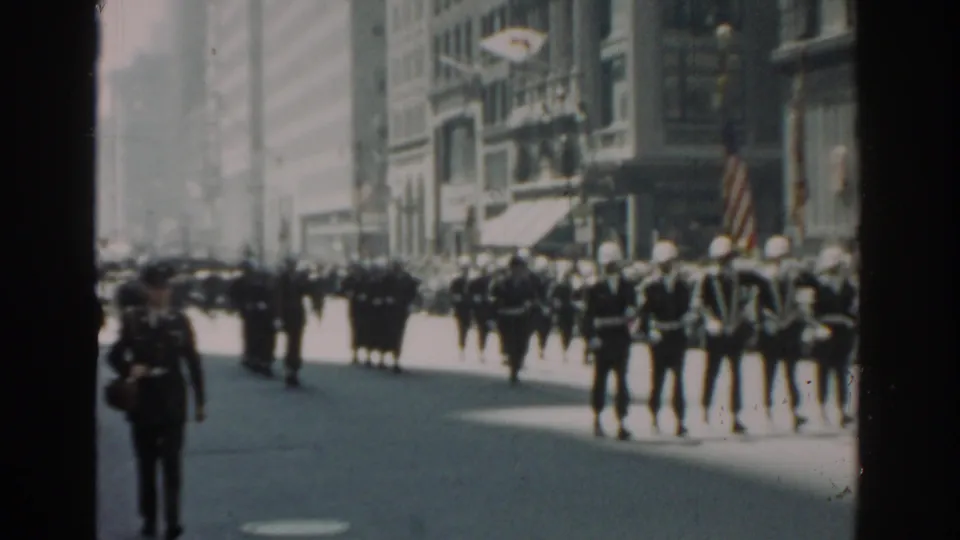 A wider handheld view shows uniformed parade participants marching across a New York City street with spectators and buildings in the background.