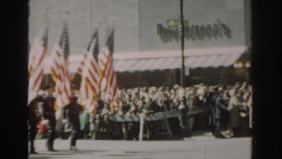 Several American flags stand near a crowded sidewalk as uniformed figures and spectators gather along the New York City parade route.