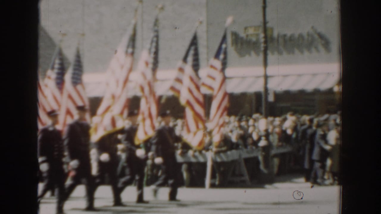 1962 New York City parade view with soldiers in dark uniforms carrying American flags past spectators and storefront signage.