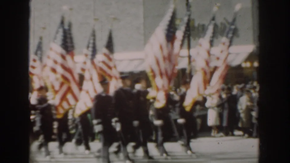 Soldiers carrying multiple American flags move through a New York City parade route as spectators watch from the sidewalk.