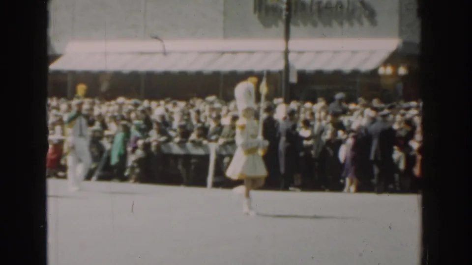 Female parade performers in white uniforms with yellow hats march along the street before a dense 1962 New York City crowd.