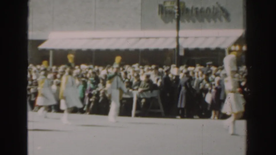 A group of women in white parade uniforms and yellow hats marches past New York City spectators in soft 8mm footage.