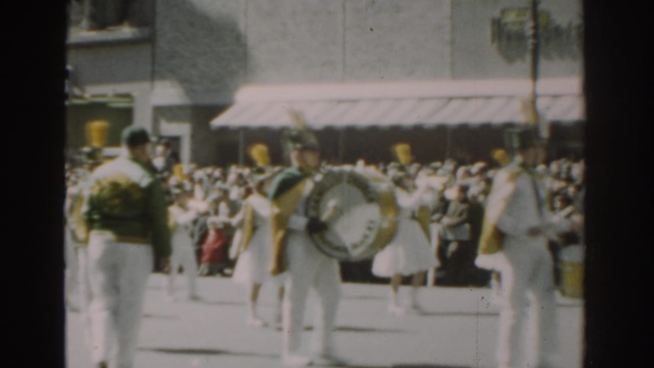 Uniformed parade drummers march past a large sidewalk crowd on a sunny city street.