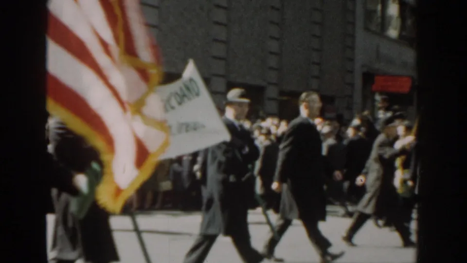 A U.S. flag moves through the 1962 New York City parade as suited participants pass a crowded sidewalk.