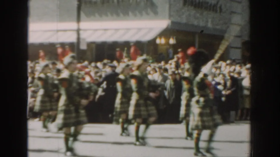 The parade continues with tartan-clad bagpipers crossing the frame before a dense New York City sidewalk crowd in 1962.