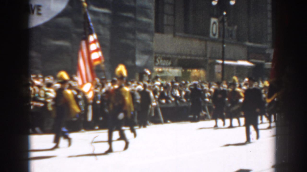 Marching band members carry an American flag past a crowd on a city street.