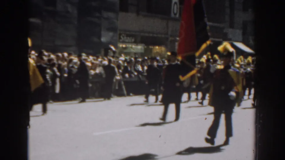 1962 New York City parade frame with banner bearers in dark uniforms and spectators watching from the sidewalk.