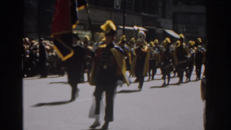 Close 8mm view of a yellow-plumed parade marcher in New York City, with other band members and spectators behind.