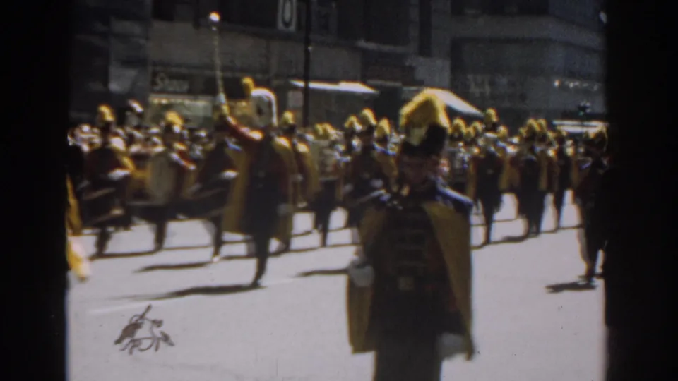 1962 New York City 8mm frame showing yellow-plumed band members marching with spectators and storefronts in the background.