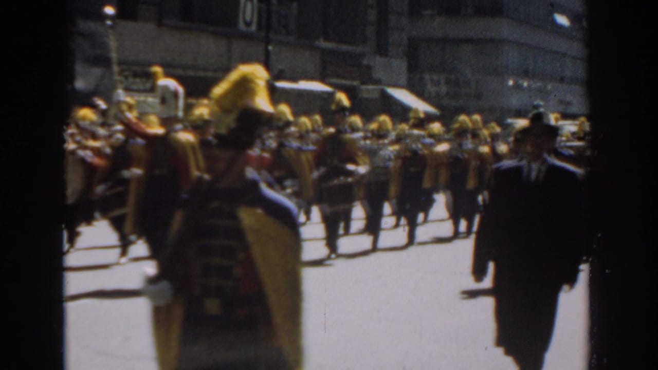 1962 New York City parade view with plumed band members, drums, spectators, and men in suits crossing the foreground.