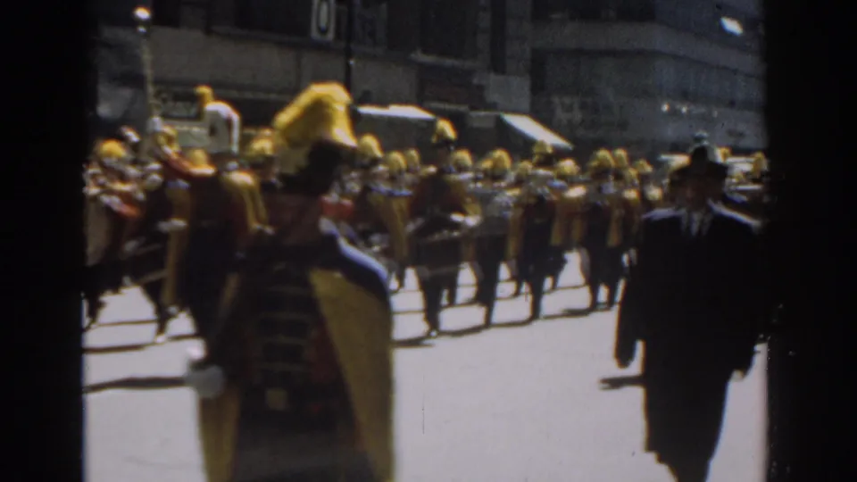 1962 New York City parade view with plumed band members, drums, spectators, and men in suits crossing the foreground.