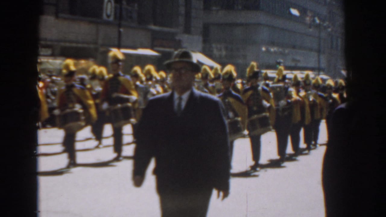 A suited man in a hat crosses before a 1962 New York City parade band wearing tall yellow plumes and carrying drums.