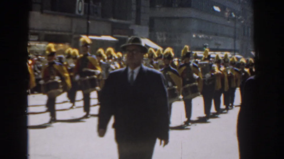 A suited man in a hat crosses before a 1962 New York City parade band wearing tall yellow plumes and carrying drums.