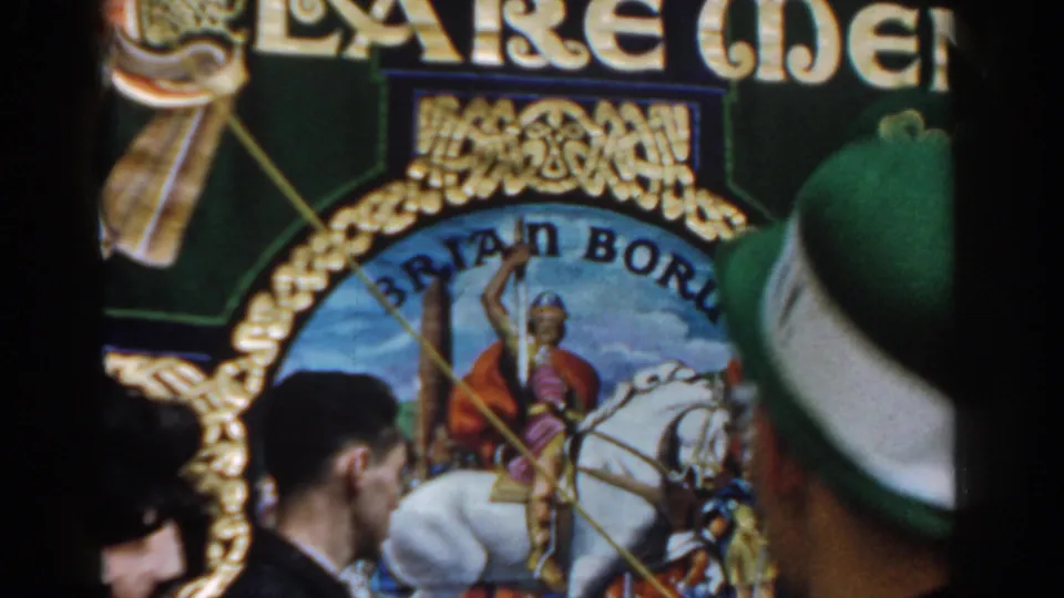 Street-side crowd view of a decorated parade banner, partially obscured by spectators and a green hat in the foreground.