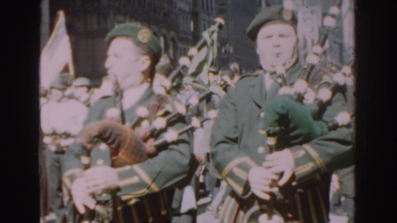 Bagpiper in a dark green uniform marches past spectators on a city street.