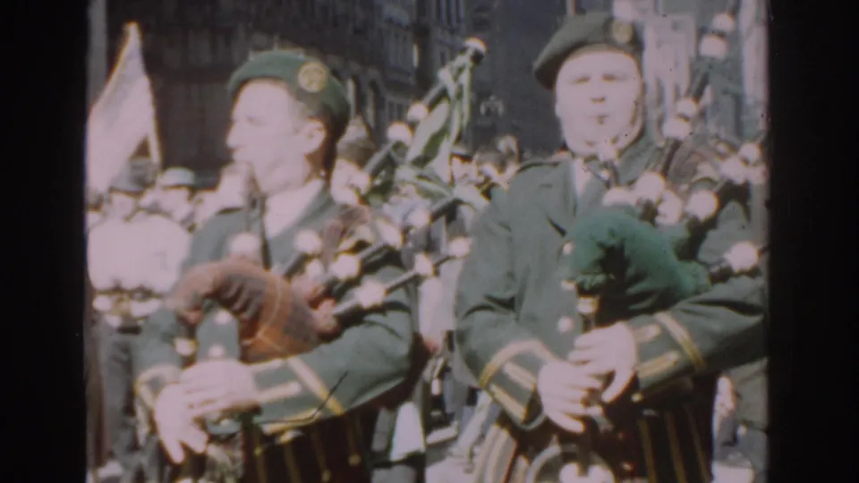 In the supplied 1962 New York City footage, a green-uniformed bagpiper marches close to the camera during a street parade.