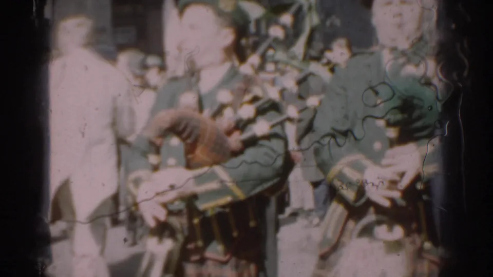 Two bagpipers in dark green uniforms and berets appear in the 1962 New York City parade footage, with spectators lining the street.
