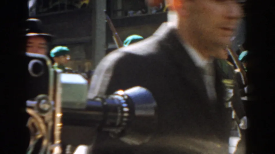 A passerby partly blocks the curbside view while uniformed parade participants and musical instruments remain visible in the 1962 New York City street scene.