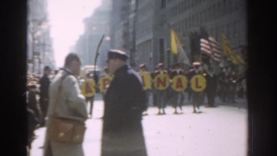 A uniformed street officer and nearby spectators stand as a parade banner and flags move along the 1962 New York City street.