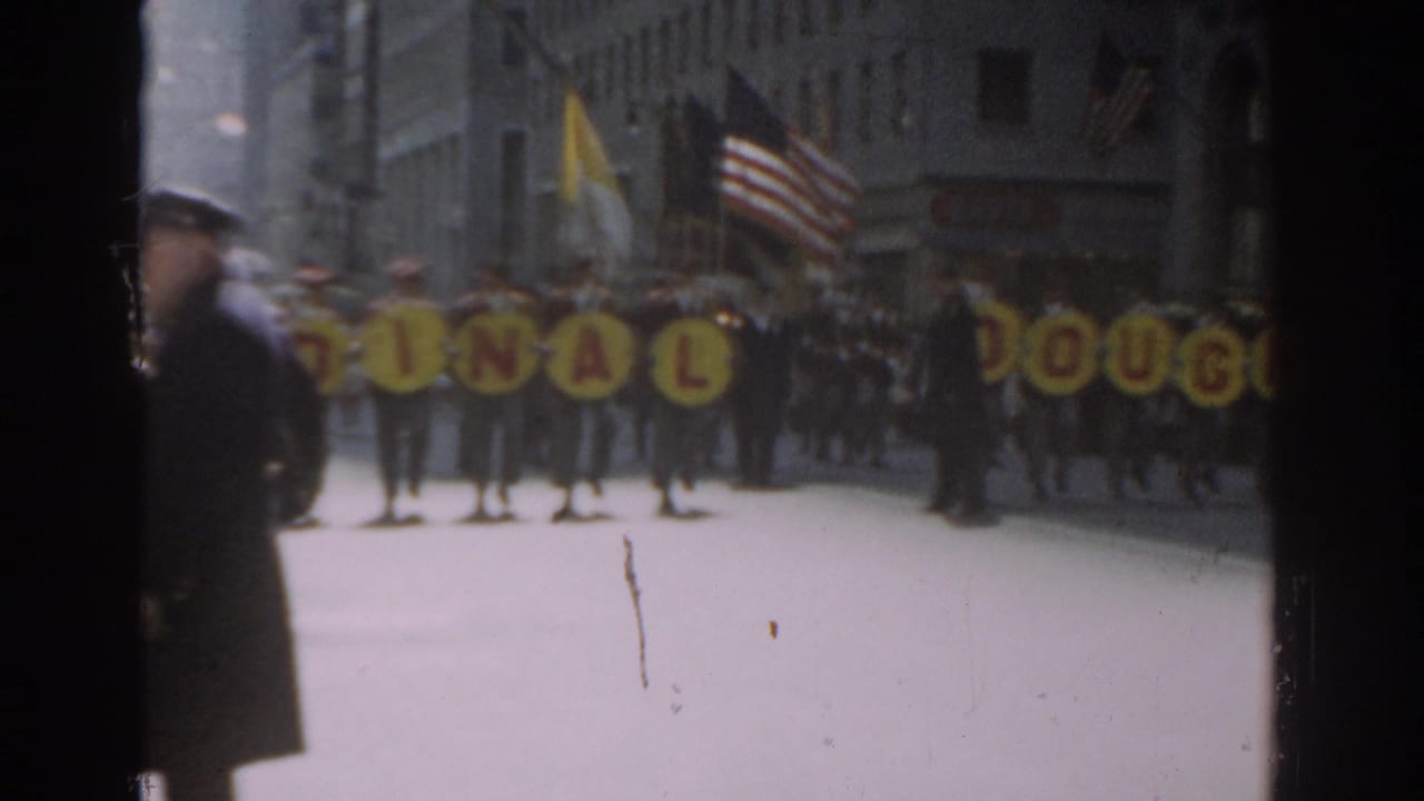 Marching band and yellow letter signs pass along a city street as a police officer stands near the curb.