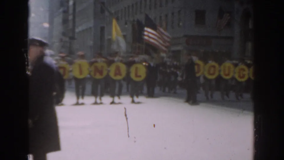 1962 New York City parade scene with band members, American flags, yellow letter signs, spectators, and a police officer at the street edge.