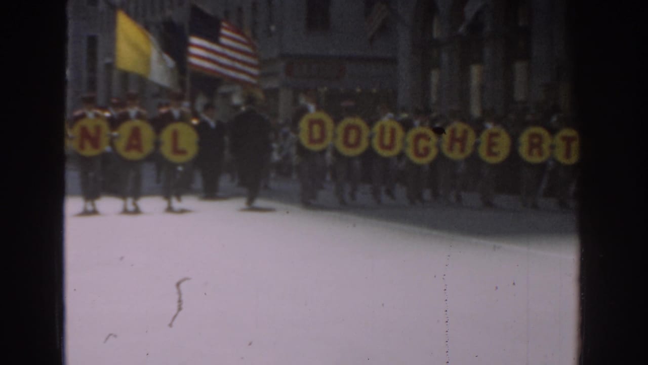 Marchers in a 1962 New York City parade carry yellow letter signs spelling part of Dinah Dougherty, with flags and spectators behind them.