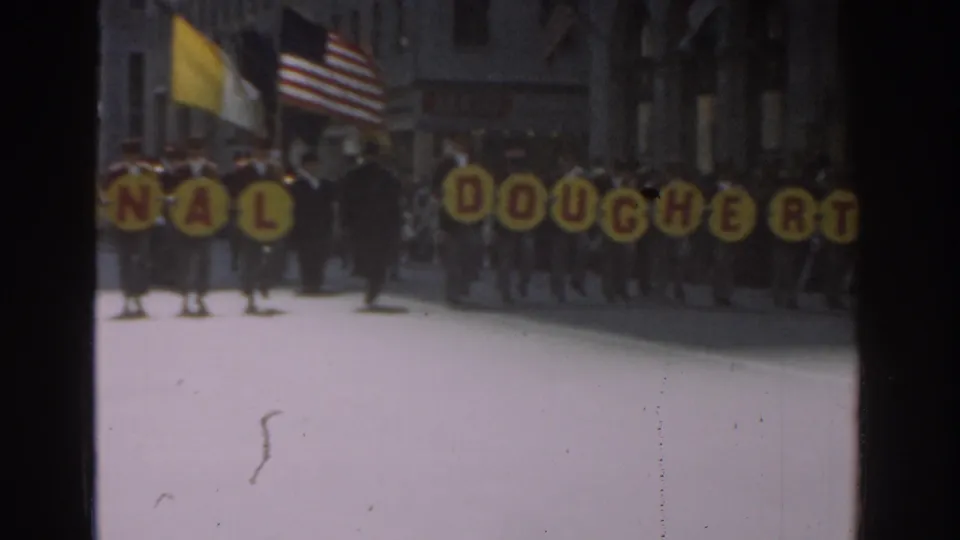 Marchers in a 1962 New York City parade carry yellow letter signs spelling part of Dinah Dougherty, with flags and spectators behind them.