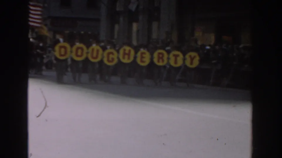 Yellow parade letters spelling Dougherty move through a dimmer 8mm frame, with marchers and spectators visible behind them in New York City.