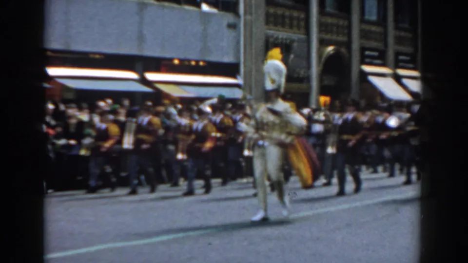 A white-uniformed parade participant stands before a marching band and spectators on a 1962 New York City street with storefront awnings.