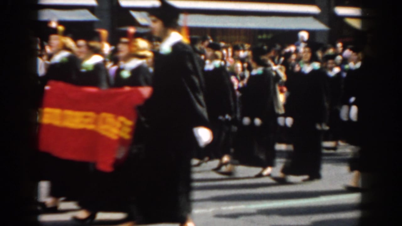 In 1962 New York City, graduates in black academic dress appear to march in a street parade with a red college banner before a crowd.