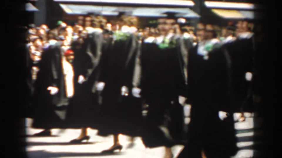 Graduates in black caps and gowns continue along a New York City street, with spectators packed behind them in bright sunlight.