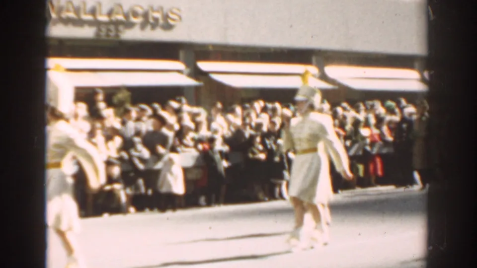 White-uniformed marchers with gold headwear perform in the street before a large New York City crowd and visible storefront signage.