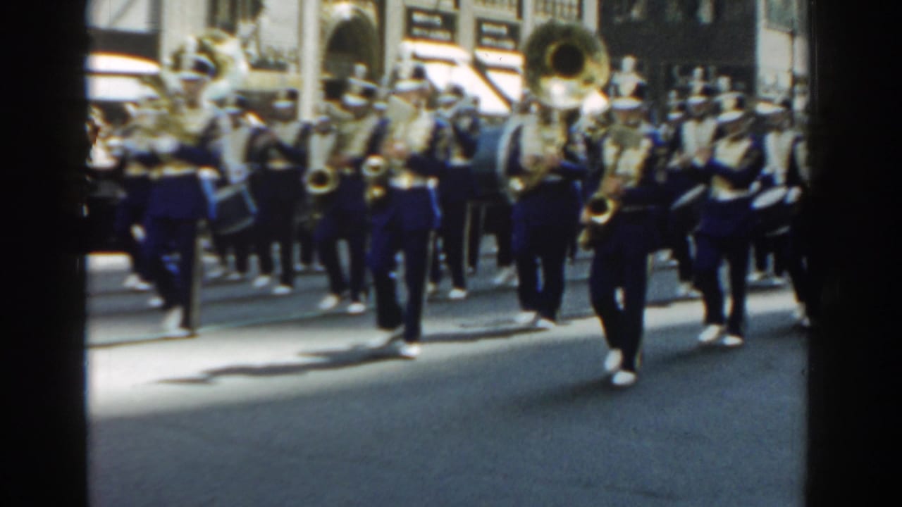 Marching band in blue-and-white uniforms advances down a city street with brass instruments.