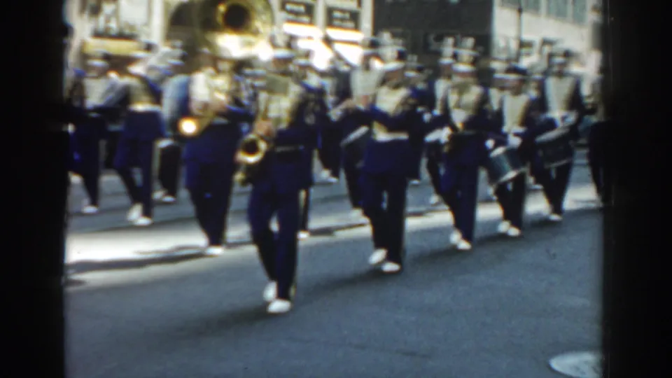 1962 New York City home-movie frame of blue-and-white uniformed band members marching in formation on a bright street.