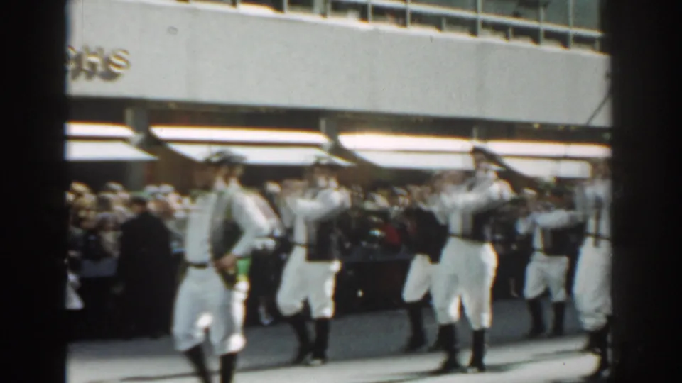 1962 New York City parade footage showing a white-uniformed marching group passing a sidewalk crowd beneath storefront awnings.
