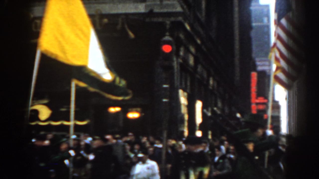 Crowded New York City street parade with flags, marchers, storefronts, and a red traffic light.