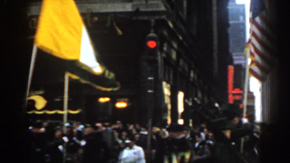1962 New York City home-movie view of a crowded street parade with flags, marchers, spectators, storefronts, and a red traffic signal.
