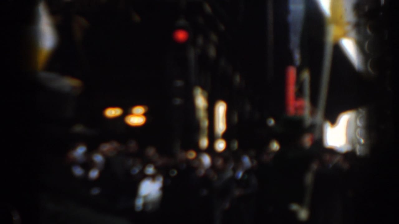 Dark city street view with blurred parade crowd, American flag, bright signs, and red traffic signal.