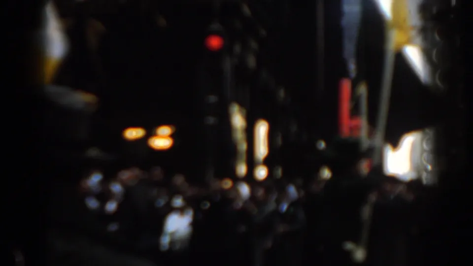 Soft 8mm view of a 1962 New York City parade crowd under tall buildings, with an American flag, bright signs, and a red traffic light.