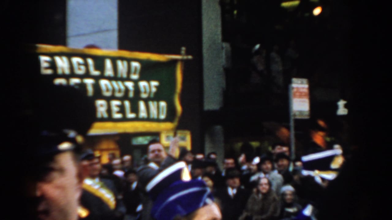 Crowd watching a parade as a green banner reading "ENGLAND GET OUT OF IRELAND" passes behind uniformed marchers.