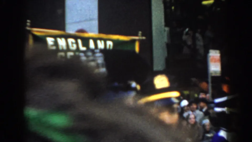 Handheld 1962 New York City parade frame with a blurred foreground passerby, spectators, and partial green banner text behind them.