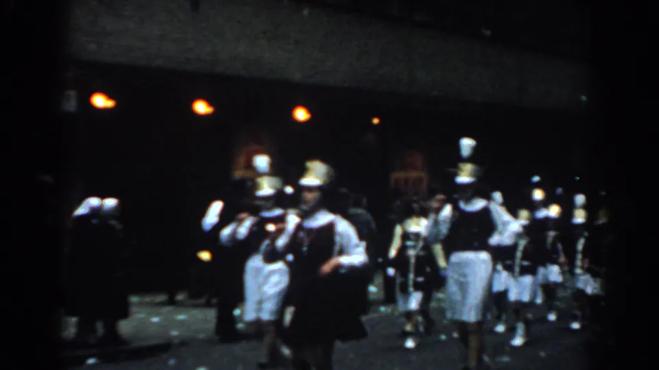 1962 New York City parade footage showing uniformed marching band members carrying instruments past a dark storefront and sidewalk spectators.