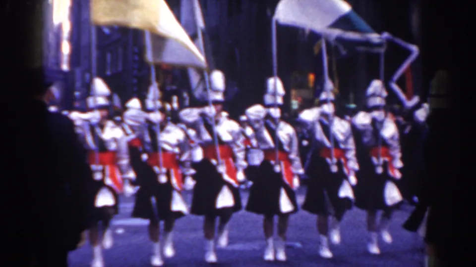 1962 New York City parade frame with white-uniformed marchers, red sashes, flags, and instruments moving in formation.