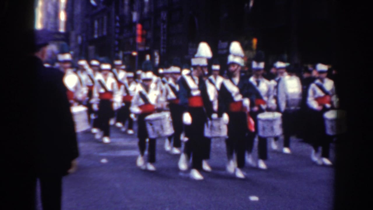 Marching band drummers in white uniforms proceed along a city street.