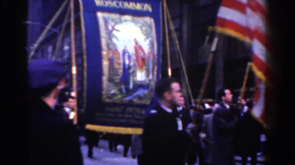 Parade participants in dark suits carry a Roscommon Society banner with religious imagery near an American flag in New York City.