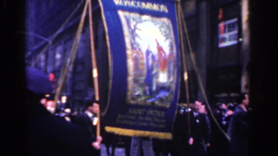 A large Roscommon Society religious banner fills the foreground as parade musicians and onlookers move through a city street.