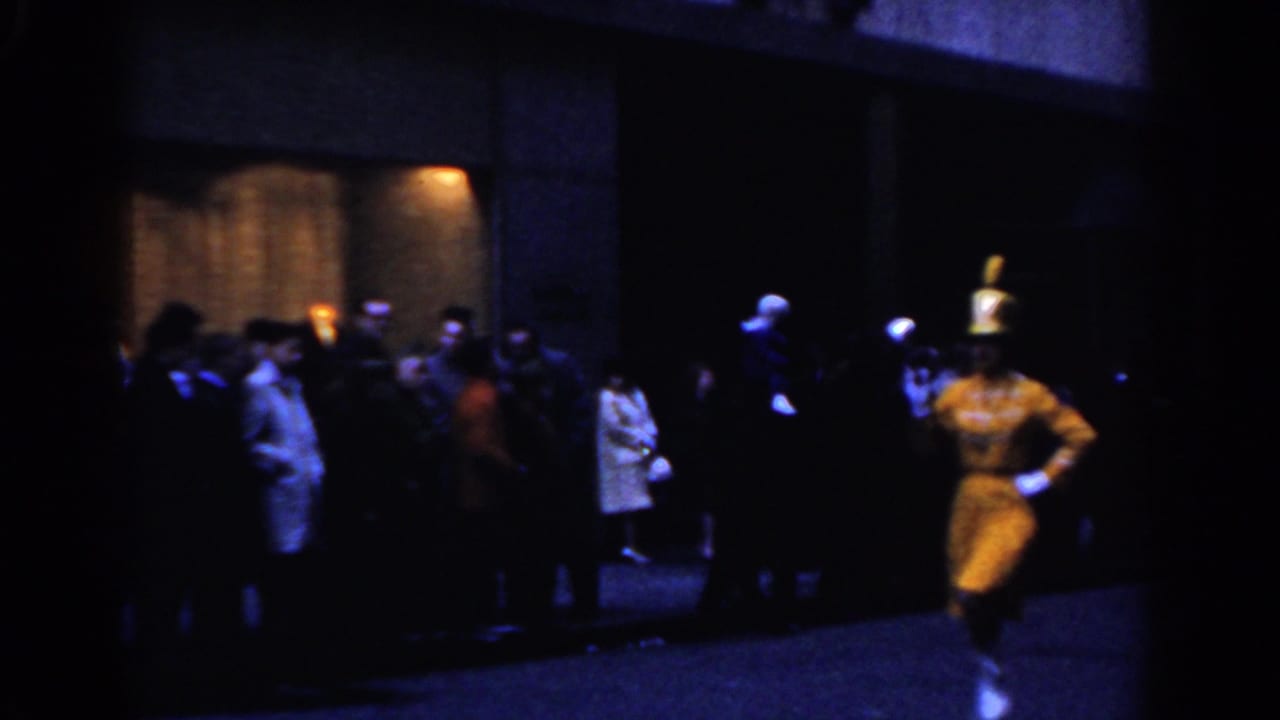 Yellow-uniformed parade performer marches past a sidewalk crowd at night.