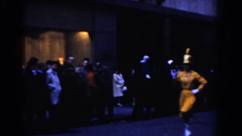 1962 New York City home-movie frame showing a yellow-uniformed marcher near spectators outside a lit building entrance at night.