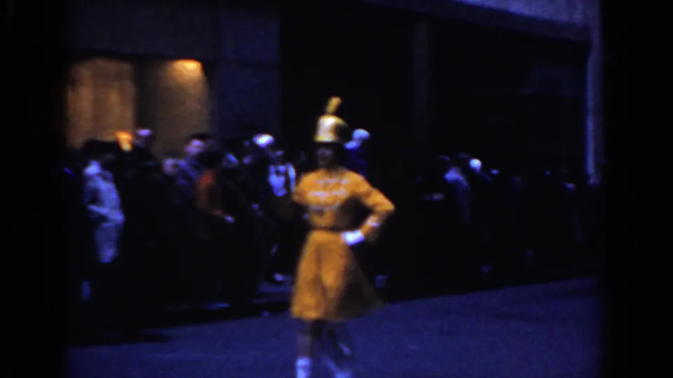 1962 New York City frame with a yellow-clad parade performer in the foreground and spectators gathered along a dark street.